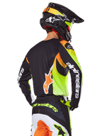 Motocrosskører set bagfra iført Alpinestars 2025 Fluid Wurx-udstyrssæt, sort trøje med neongul og orange grafik, matchende bukser med hvide og orange paneler, sort hjelm med neon-detaljer, tydelig Alpinestars-branding på ærmer og bagpå