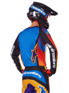 Motocrossrytter set bagfra iført Alpinestars 2025 Fluid Wurx-udstyrssæt, flerfarvet trøje med blå ryg og sorte ærmer med hvide logoer, matchende orange og blå bukser, sort og blå hjelm med orange briller, koordineret off-road raceroutfit.