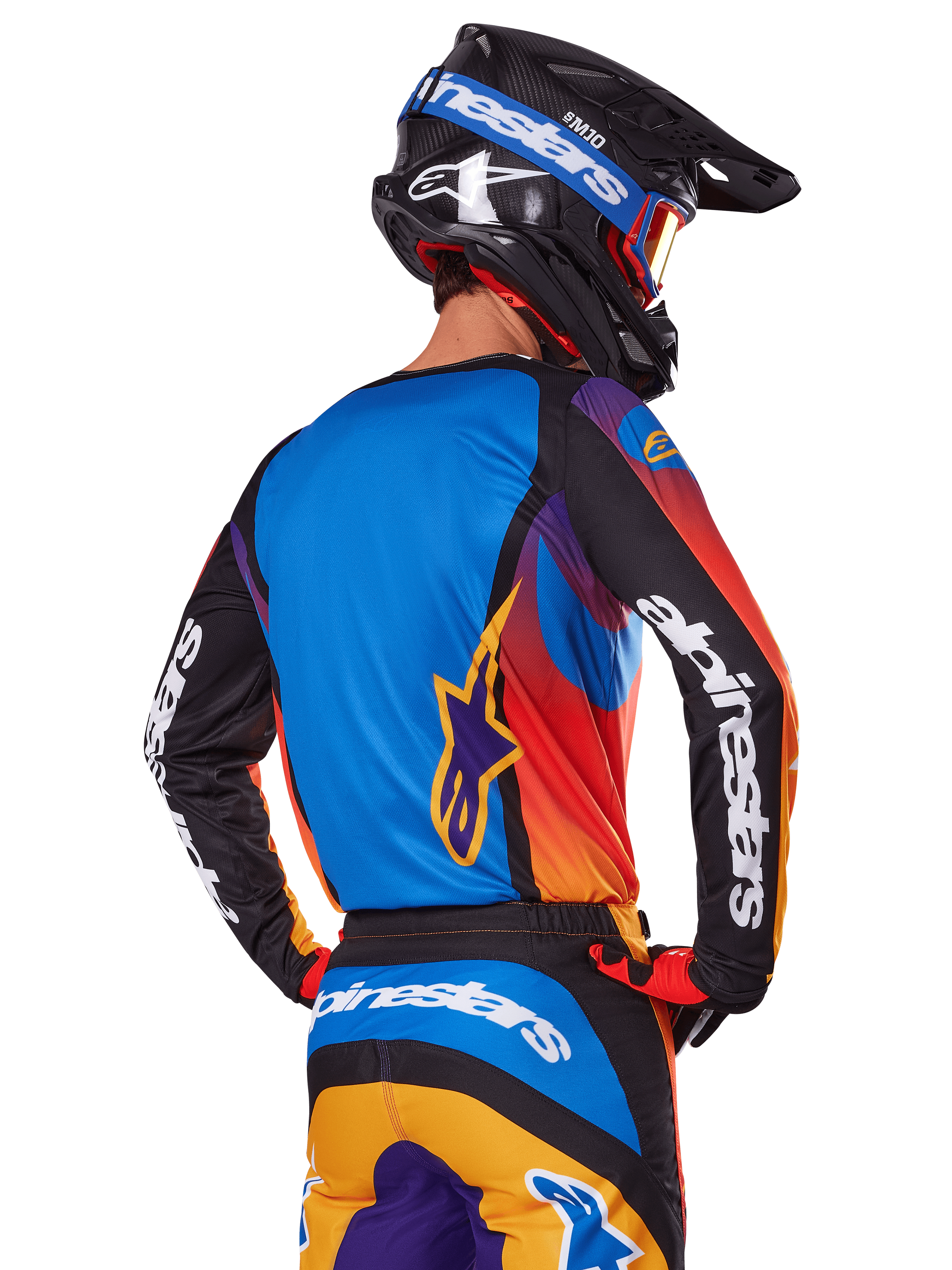 Motocrossrytter set bagfra iført Alpinestars 2025 Fluid Wurx-udstyrssæt, flerfarvet trøje med blå ryg og sorte ærmer med hvide logoer, matchende orange og blå bukser, sort og blå hjelm med orange briller, koordineret off-road raceroutfit.