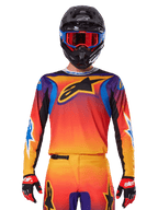 Alpinestars 2025 Fluid Wurx trøje, multicolor motocross-trøje med orange og rød farveovergang og lilla detaljer, vist med matchende udstyr, sort hjelm med blå og røde goggles og røde handsker, med et stort sort stjernelogo på brystet.