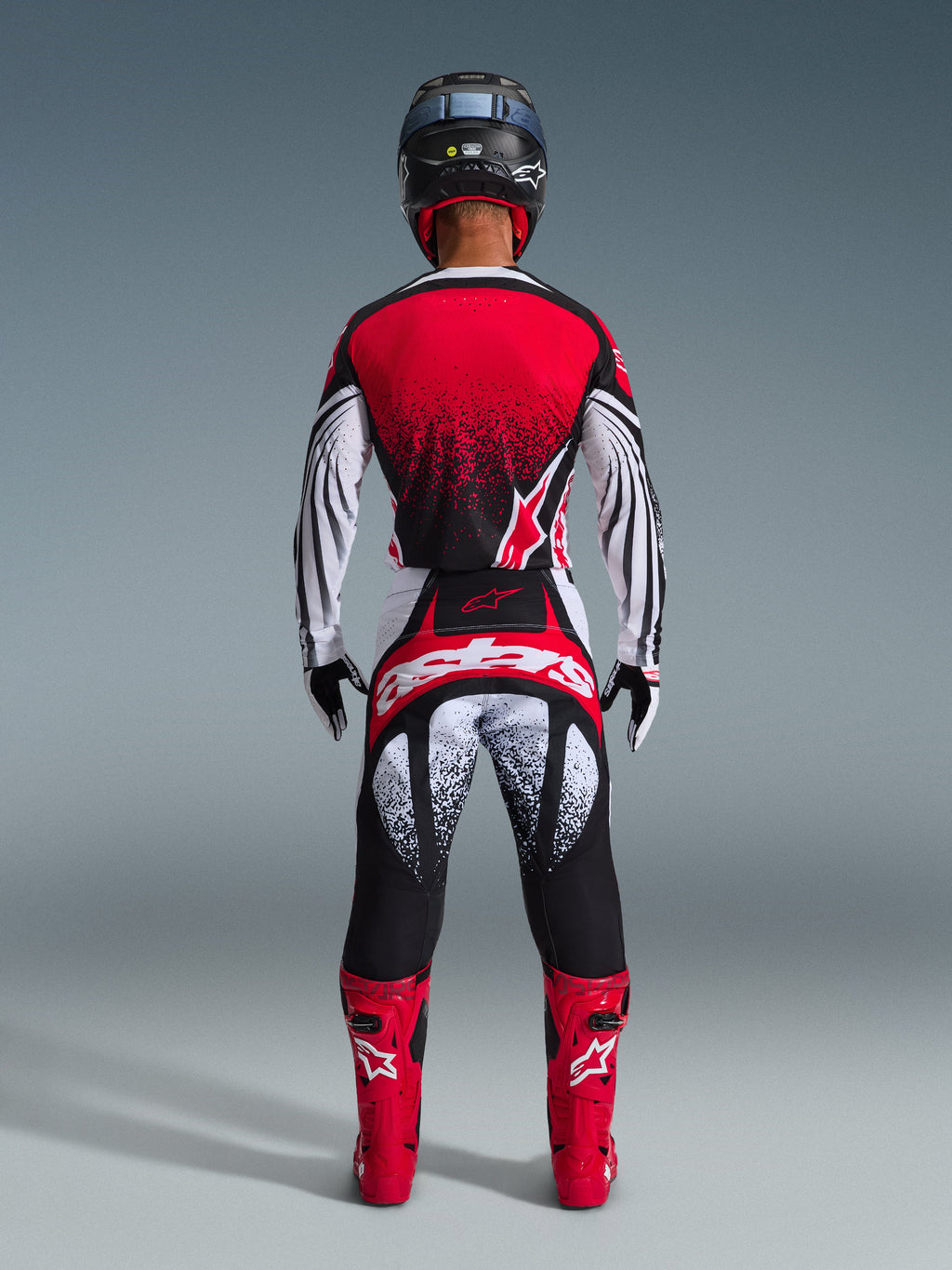 Motocrosskører set bagfra iført Alpinestars 2026 Techstar Nomur-udstyrssæt, rød og hvid jersey med sort nistret gradient, matchende hvide og røde bukser med sorte detaljer, klare røde støvler og sort hjelm, koordineret off-road racingudstyr.