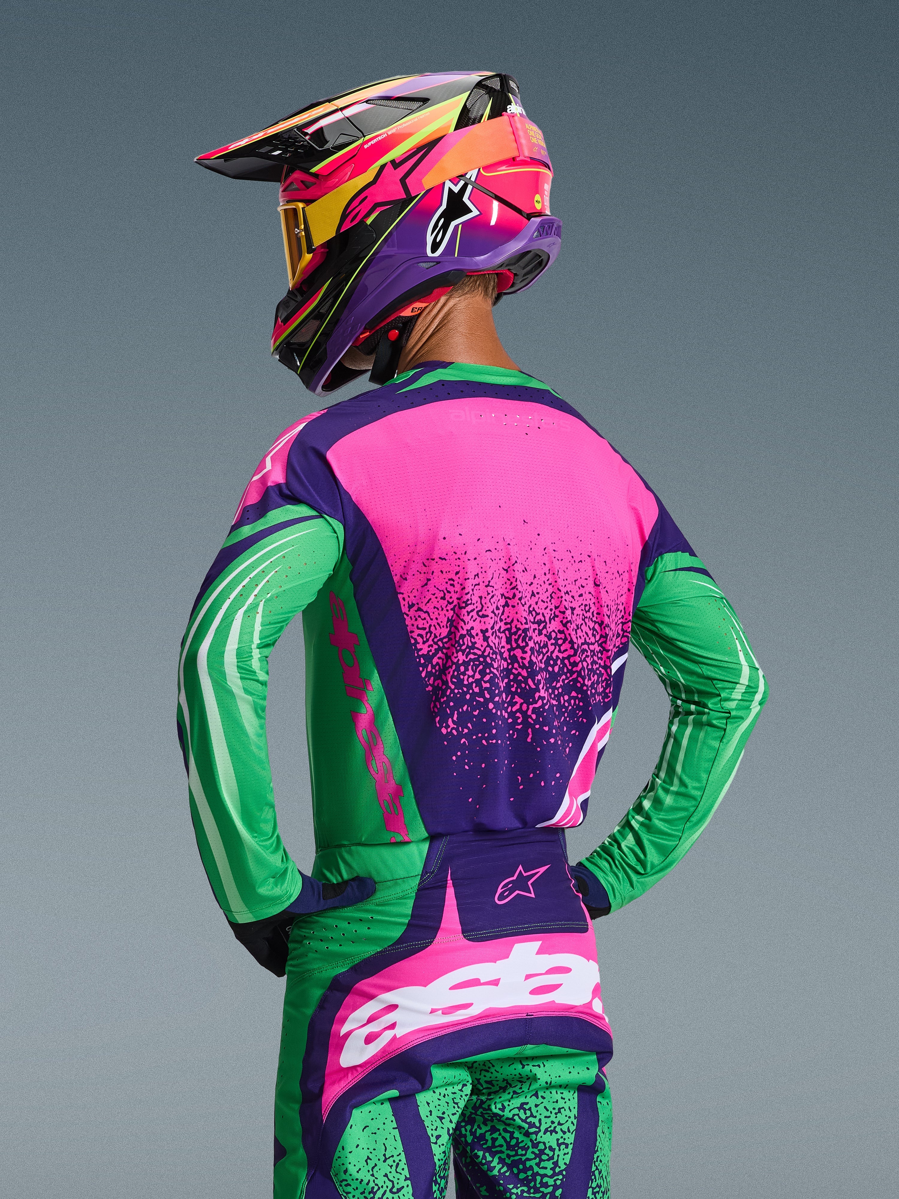 Motocross-kører set bagfra iført Alpinestars 2026 Techstar Nomur-udstyrssæt, mørkelilla, grøn og pink fluo trøje og matchende bukser, med markant grafik og en atletisk pasform, kombineret med en flerfarvet hjelm og gule goggles.