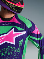 Nærbillede af Alpinestars 2026 Techstar Nomur Jersey, mørkelilla med grønne og pink fluo-detaljer, med et stort pink stjernelogo på brystet og laserskårne perforeringer på skulderen, båret af en kører med en matchende lilla og pink motocrosshjelm.