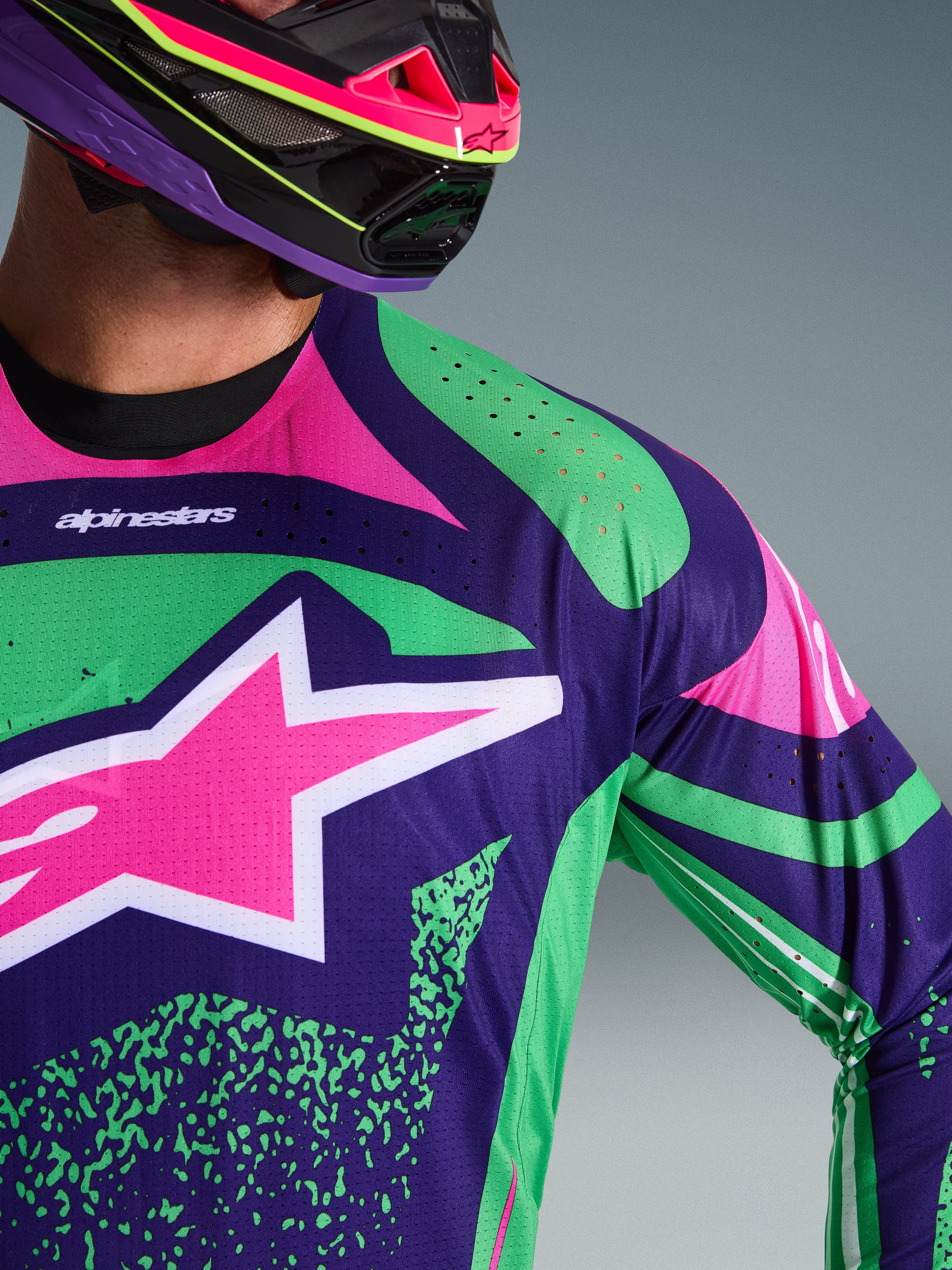 Nærbillede af Alpinestars 2026 Techstar Nomur Jersey, mørkelilla med grønne og pink fluo-detaljer, med et stort pink stjernelogo på brystet og laserskårne perforeringer på skulderen, båret af en kører med en matchende lilla og pink motocrosshjelm.