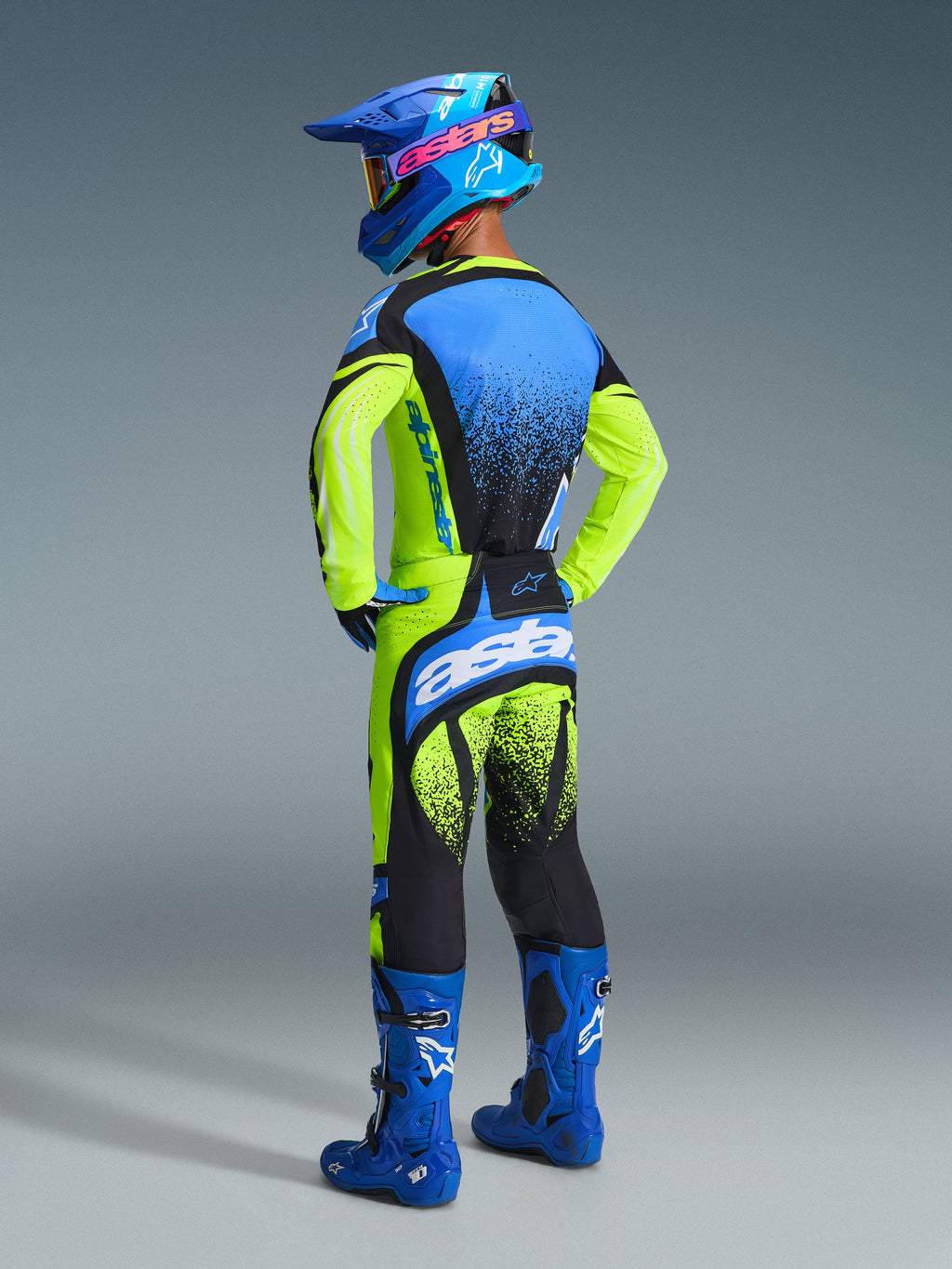 Alpinestars 2026 Techstar Nomur-udstyrssæt, motocrosskører set bagfra, fluorescerende gul og blå trøje med sorte nistrede detaljer, matchende bukser med markant hvid branding, blå racingstøvler og blå hjelm, komplet professionelt raceroutfit til terrænkørsel