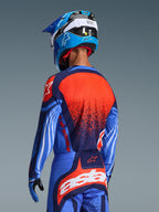 Motocross-rytter set bagfra iført Alpinestars 2026 Techstar Nomur-trøje og matchende bukser, mørkeblå og UCLA-blå med livlige orange detaljer, med en stor stjerne-grafik og laserskåret ventilation på trøjen, kombineret med en blå og hvid hjelm for et komplet off-road racersæt