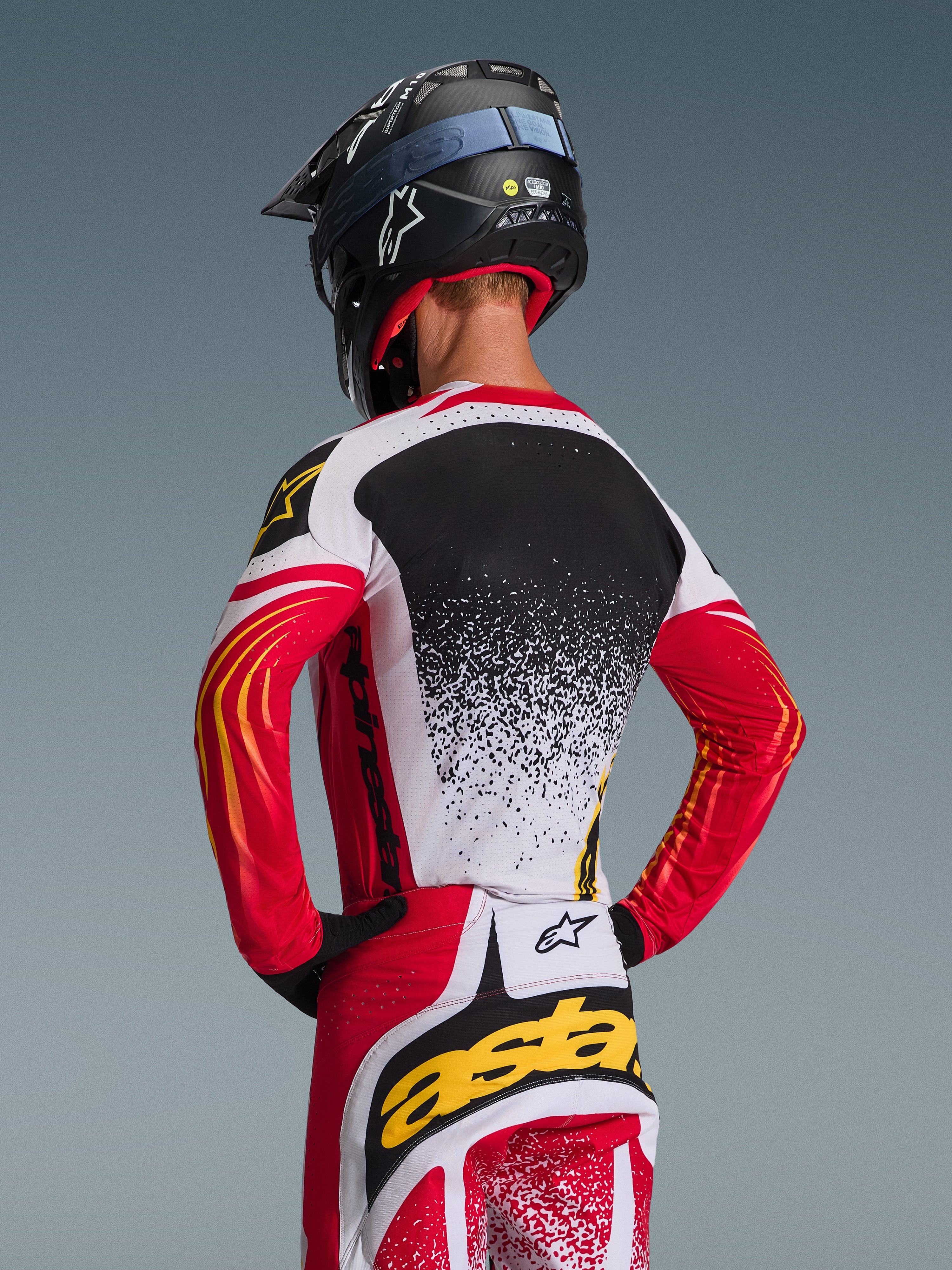 Alpinestars 2026 Techstar Nomur udstyrssæt, motocross trøje og bukser, rød, hvid og sort med gule detaljer, set bagfra af kører iført sort hjelm, performance racerudstyr med splatter-grafik og markant branding