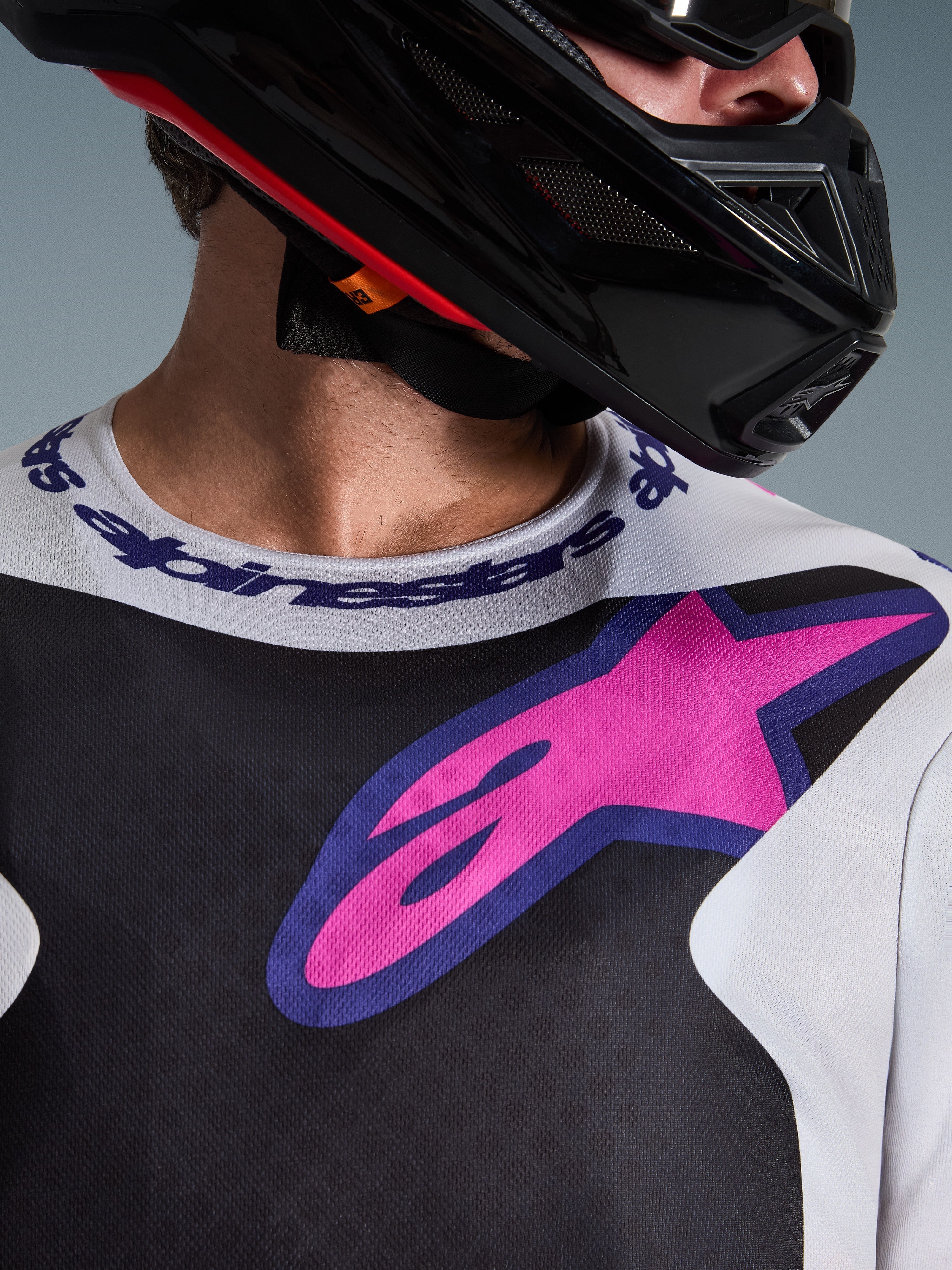 Alpinestars 2026 Fluid Grid trøje, motocross-trøje, sort, lysegrå og lilla, nærbillede af bryst- og halsområde med stort pink stjernelogo, sportskrave med branding, båret med sort hjelm, performance off-road-udstyr