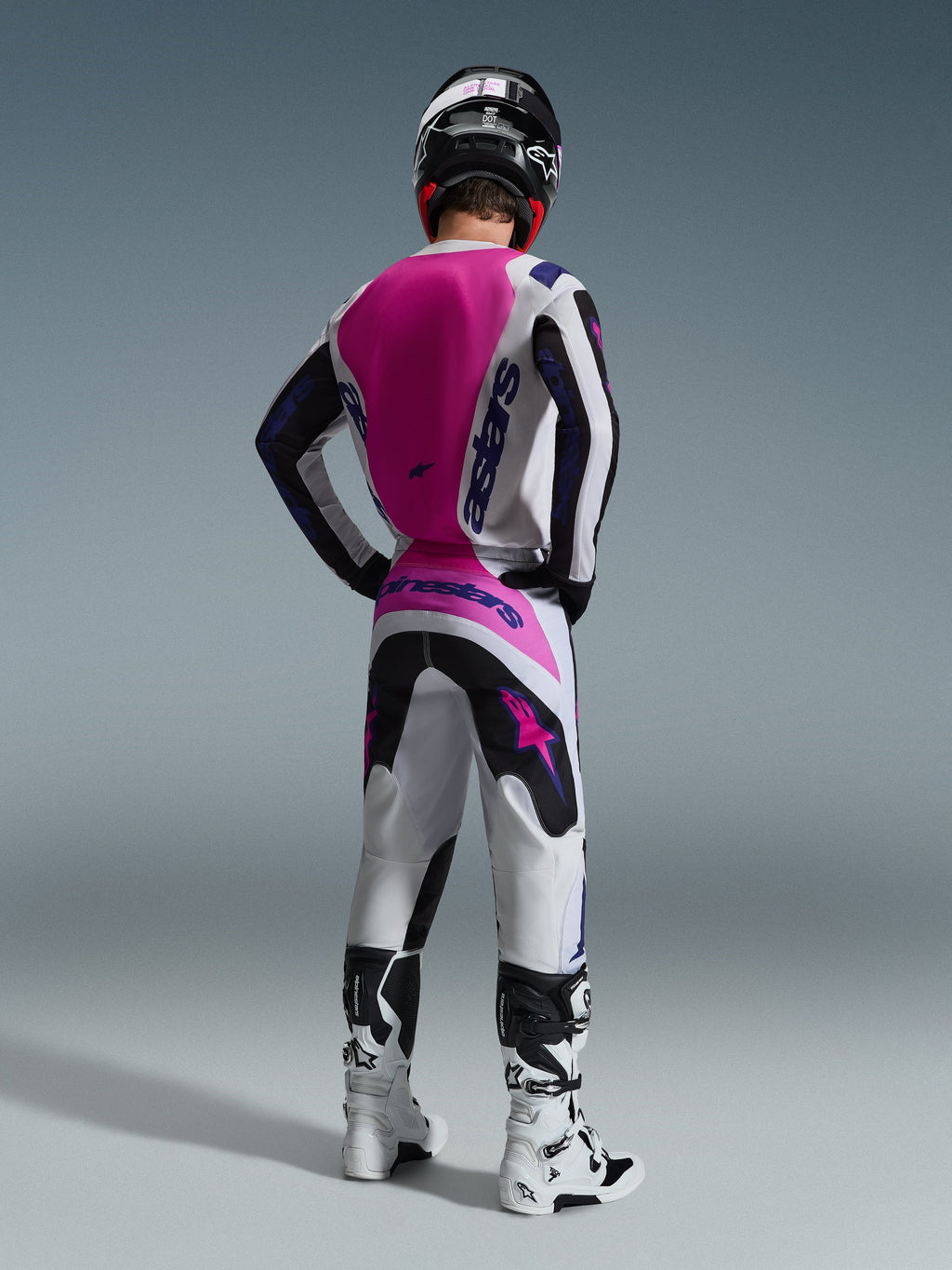 Motocross-kører set bagfra iført Alpinestars 2026 Fluid Grid trøje, hvid trøje med stort pink rygpanel og lilla branding, matchende hvide og sorte bukser med pink detaljer, hvide støvler og sort hjelm, koordineret off-road racerudstyr