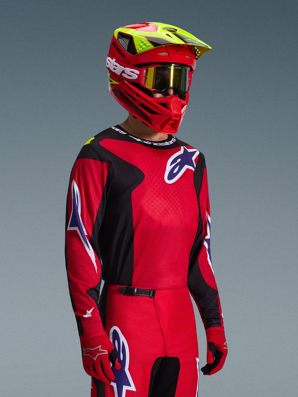 Motocross-kører iført Alpinestars 2026 Fluid Grid-trøje i rød, sort og lilla, koordineret med matchende motocross-bukser, en rød hjelm med neongule detaljer, guld-goggles og røde handsker, komplet off-road raceroutfit.