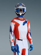 Motocross-kører iført Alpinestars 2026 Fluid Grid-udstyrssæt, orange og hvid jersey med blå stjernelogoer, hvide og orange bukser med blå detaljer, blå handsker, blå og hvid hjelm med blå goggles, koordineret atletisk racerudstyr til off-road-performance