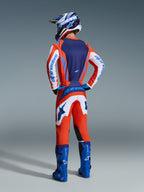 Motocrosskører set bagfra iført 2026 Fluid Grid Jersey og bukser i Orange og UCLA Blue, med hvide ærmer og blåt Alpinestars-branding, klare blå motocrossstøvler og en matchende hjelm, komplet udstyrssæt til terrænkørsel.