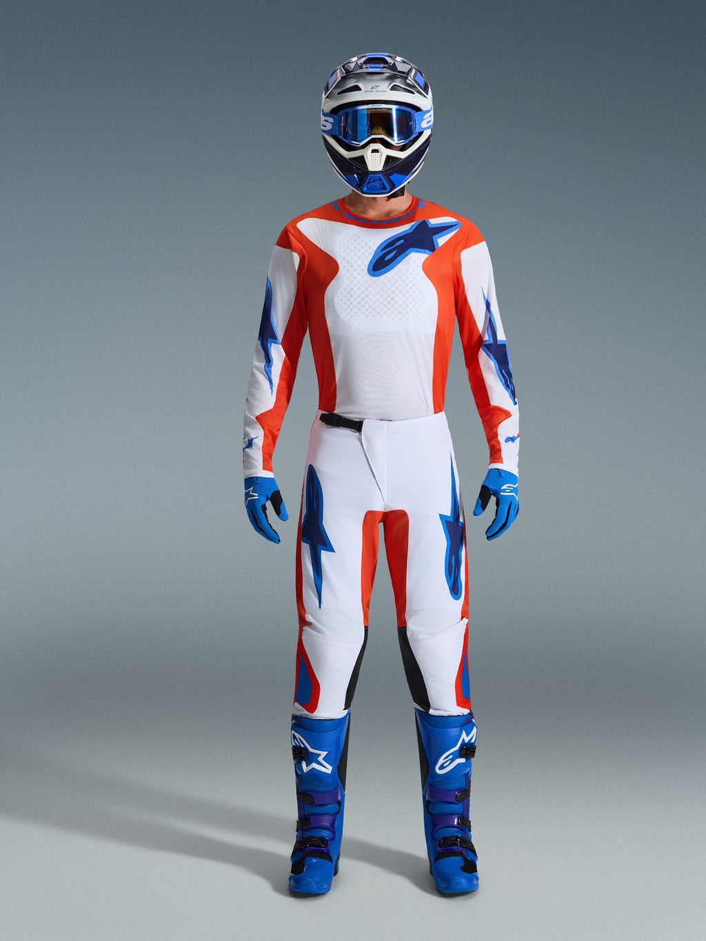 Alpinestars 2026 Fluid Grid udstyrssæt, motocross-rytter i fuldt udstyr, hvid trøje og bukser med orange og UCLA blå detaljer, matchende blå og hvid hjelm med blå motocross-briller, blå handsker og blå motocross-støvler, koordineret raceroutfit til terrænkørsel