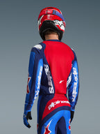 Set bagfra af en motocross-kører iført Alpinestars 2026 Fluid Grid-trøjen i blå og rød, med markant hvid branding på ærmerne og ryggen, kombineret med matchende motocross-bukser og en rød hjelm for et komplet off-road racerlook