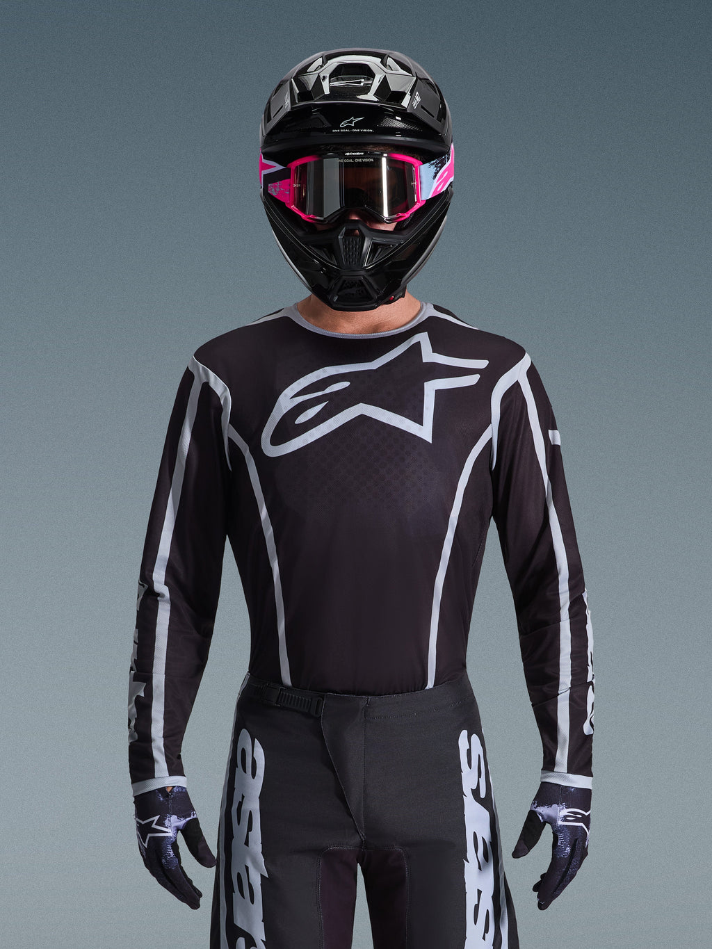 Alpinestars 2026 Fluid Apex trøje, motocross-trøje, sort og grå, med et stort hvidt Astars-logo på brystet og grå grafiske detaljer på ærmerne, båret af en kører med lyserøde goggles og en sort hjelm, koordineret off-road-racerudstyr