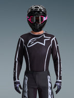 Alpinestars 2026 Fluid Apex trøje, motocross-trøje, sort og grå, med et stort hvidt Astars-logo på brystet og grå grafiske detaljer på ærmerne, båret af en kører med lyserøde goggles og en sort hjelm, koordineret off-road-racerudstyr