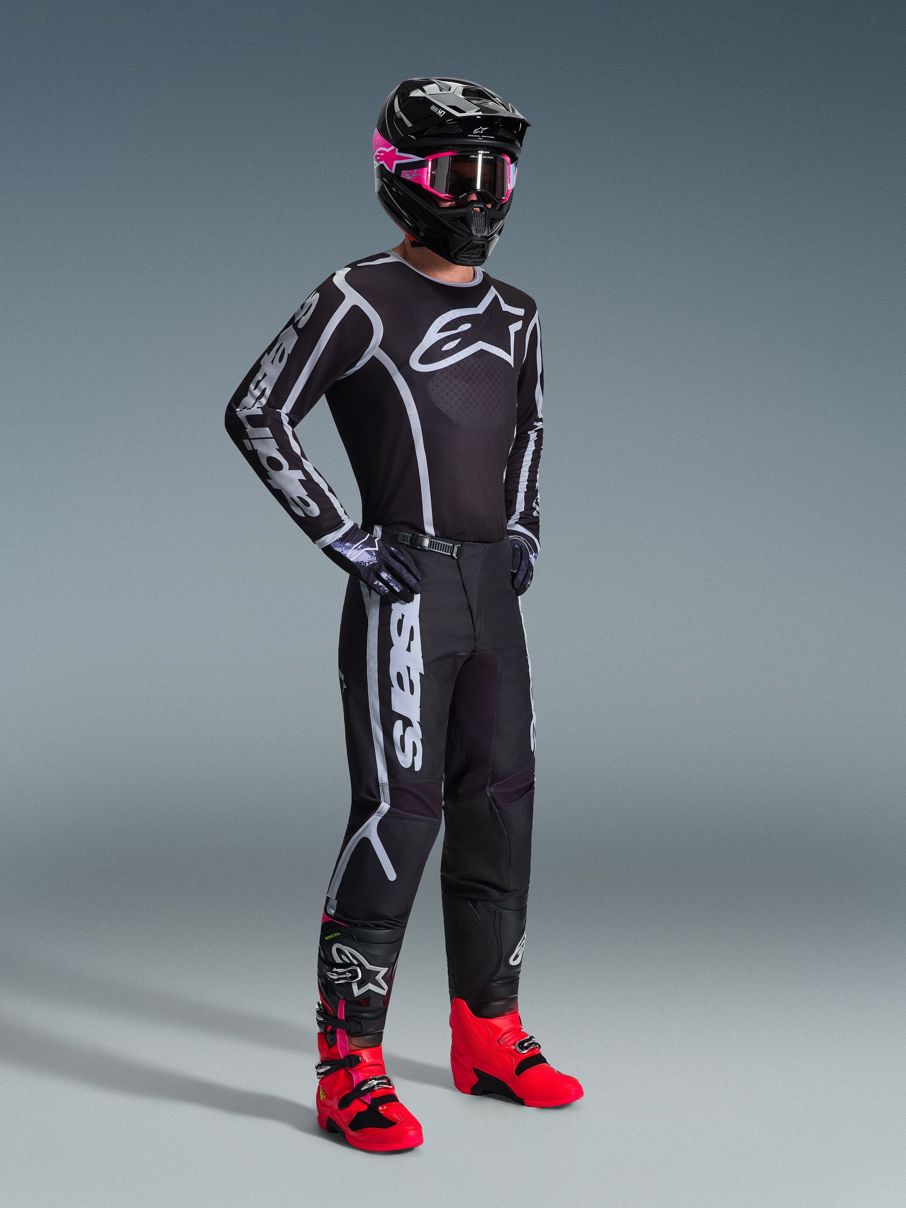 Motocross-kører iført 2026 Fluid Apex trøje, sort og grå, matchende bukser, sort hjelm med lyserøde goggles og knaldrøde støvler, markant hvidt Alpinestars-branding på bryst og ærmer, koordineret professionelt off-road raceroutfit.