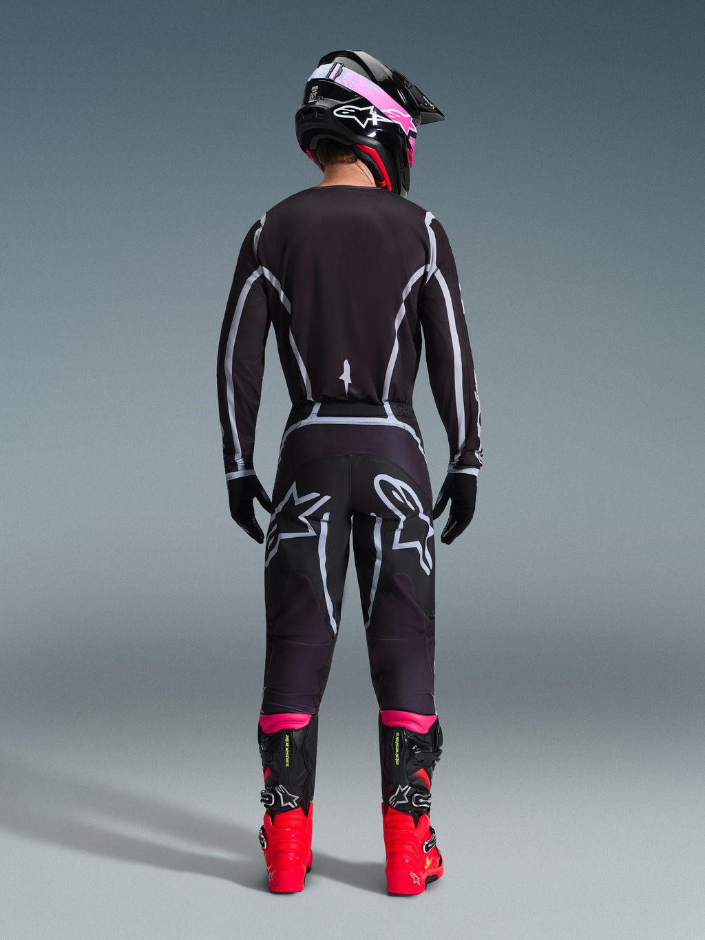 Motocross-kører set bagfra iført Alpinestars 2026 Fluid Apex trøje og matchende bukser i sort og grå, med hvide geometriske designlinjer og stjernelogoer på bagdelen, suppleret med en sort og pink hjelm og røde og sorte racingstøvler.