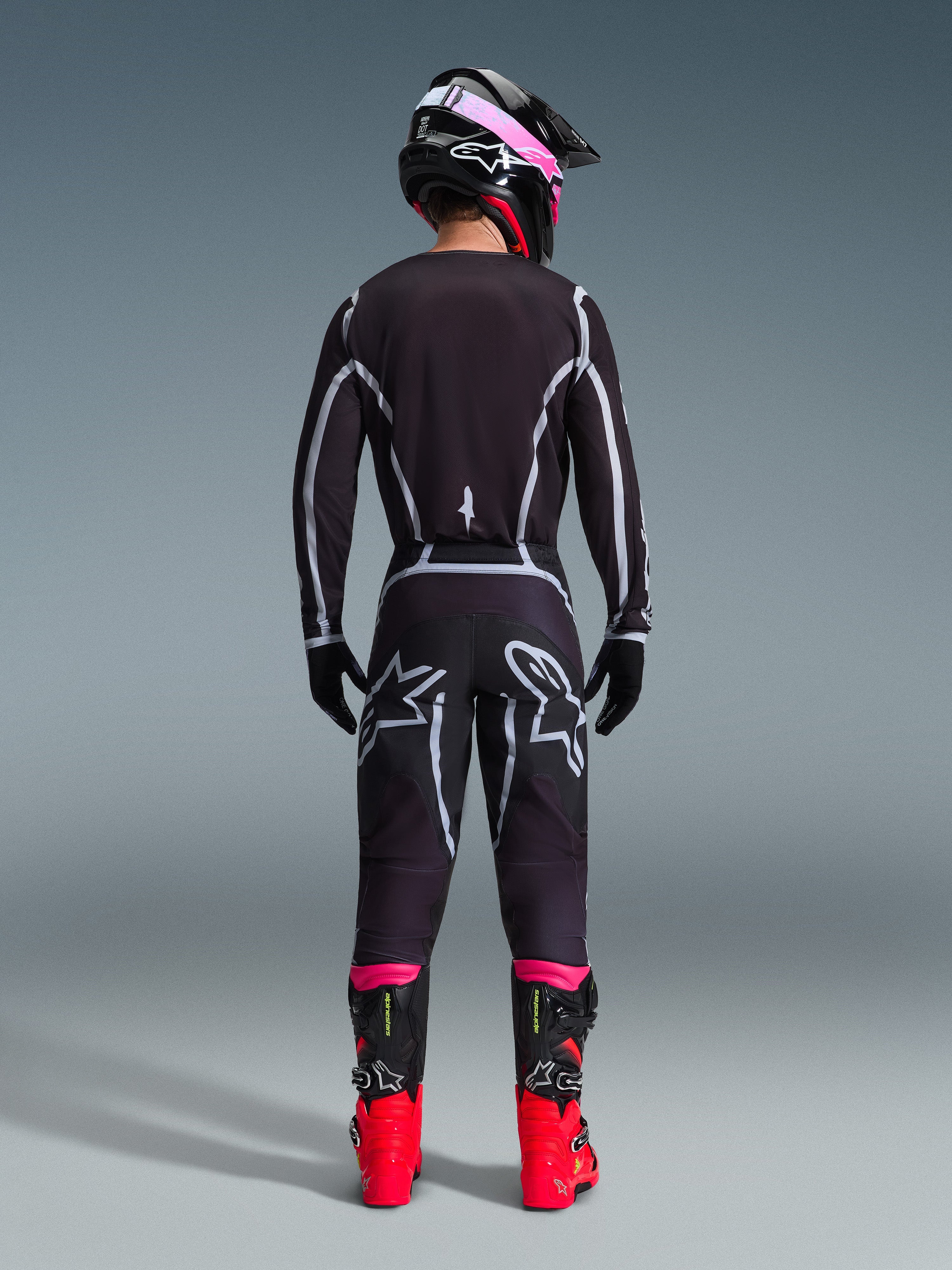 Motocross-kører set bagfra iført Alpinestars 2026 Fluid Apex trøje og matchende bukser i sort og grå, med hvide geometriske designlinjer og stjernelogoer på bagdelen, suppleret med en sort og pink hjelm og røde og sorte racingstøvler.