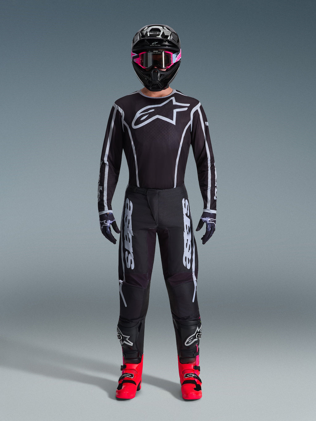 Motocrosskører iført Alpinestars 2026 Fluid Apex Jersey, sort og grå, stort hvidt Astars-logo på brystet, hvide geometriske linjer, matchende sorte bukser, sort hjelm med lyserøde goggles og panglyserøde støvler, komplet motocross-raceroutfit