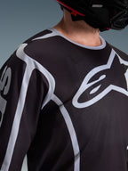 Nærbillede af en kører iført Alpinestars 2026 Fluid Apex Jersey i sort og grå, der fremviser det store hvide Astars-logo på brystet og den tekniske mesh-konstruktion, kombineret med en sort motocrosshjelm.