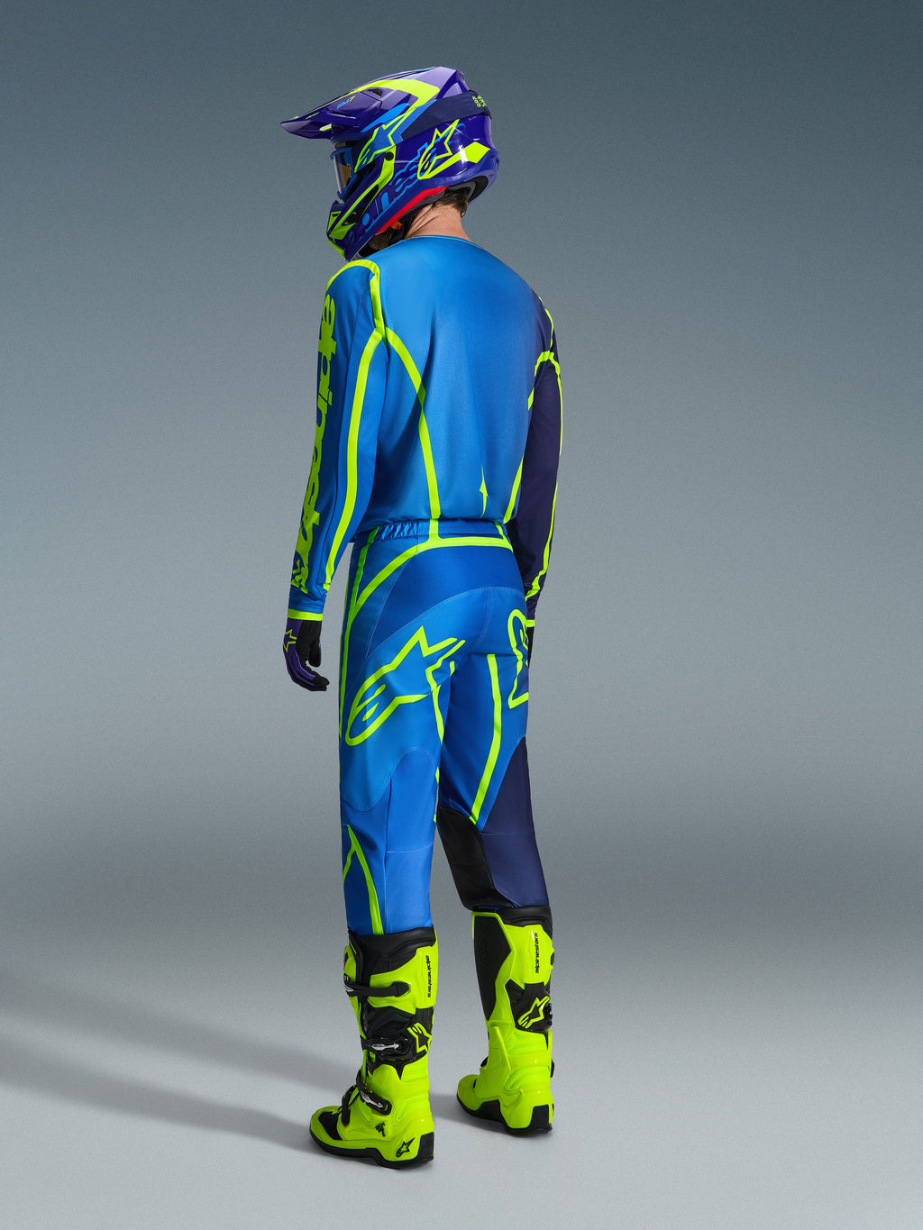 Motocross-kører set bagfra iført Alpinestars 2026 Fluid Apex Jersey i Blue Yellow Fluo, matchende blå og fluorescerende gule bukser, skarpt gule støvler og en koordineret hjelm, professionelt off-road racerudstyr