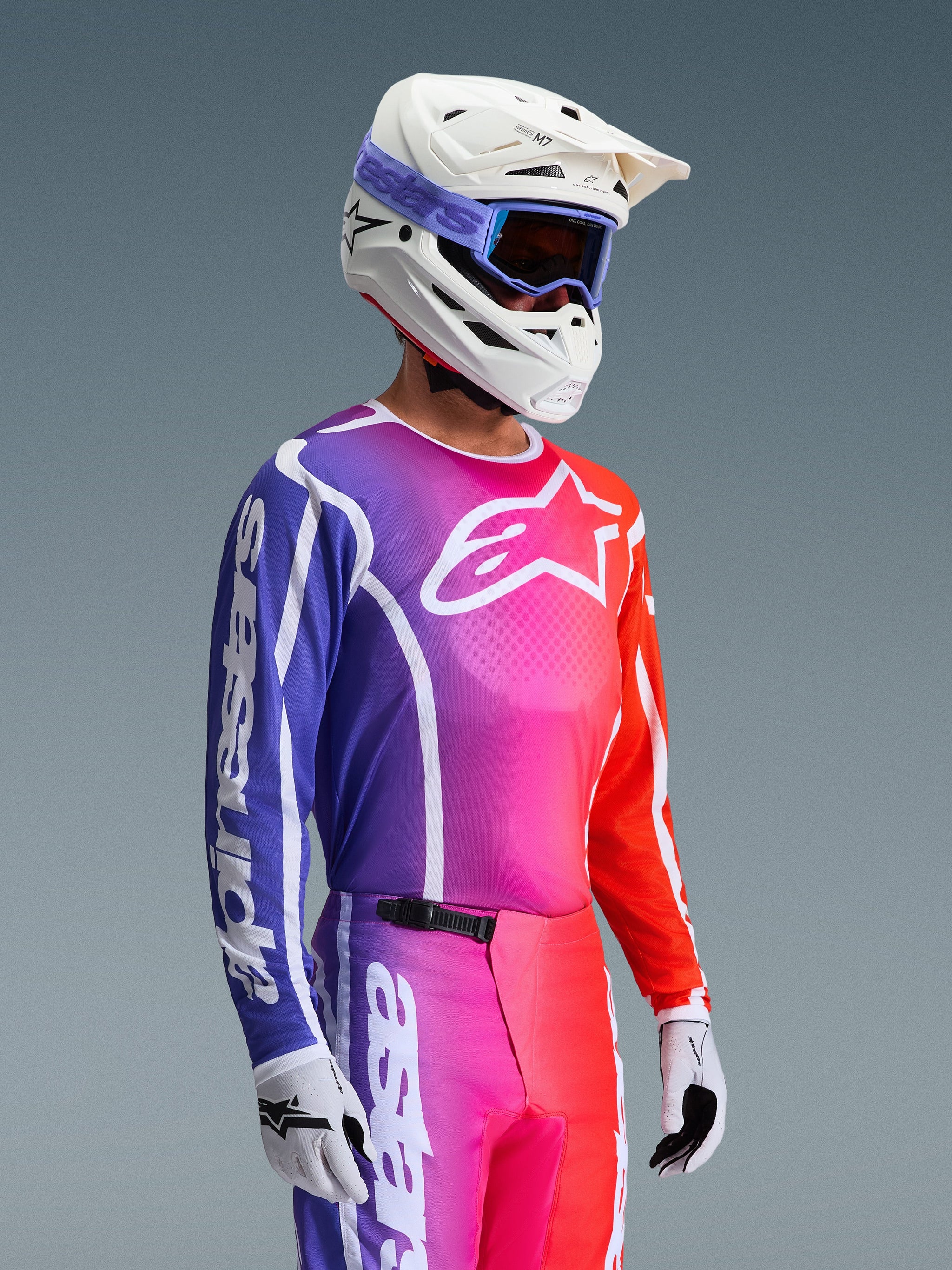 Alpinestars 2026 Fluid Apex Jersey, motocross-trøje, levende lilla, lyserød og orange farveovergang, båret med matchende bukser, hvid hjelm med blå goggles og hvide handsker, atletisk design til off-road performance og mobilitet