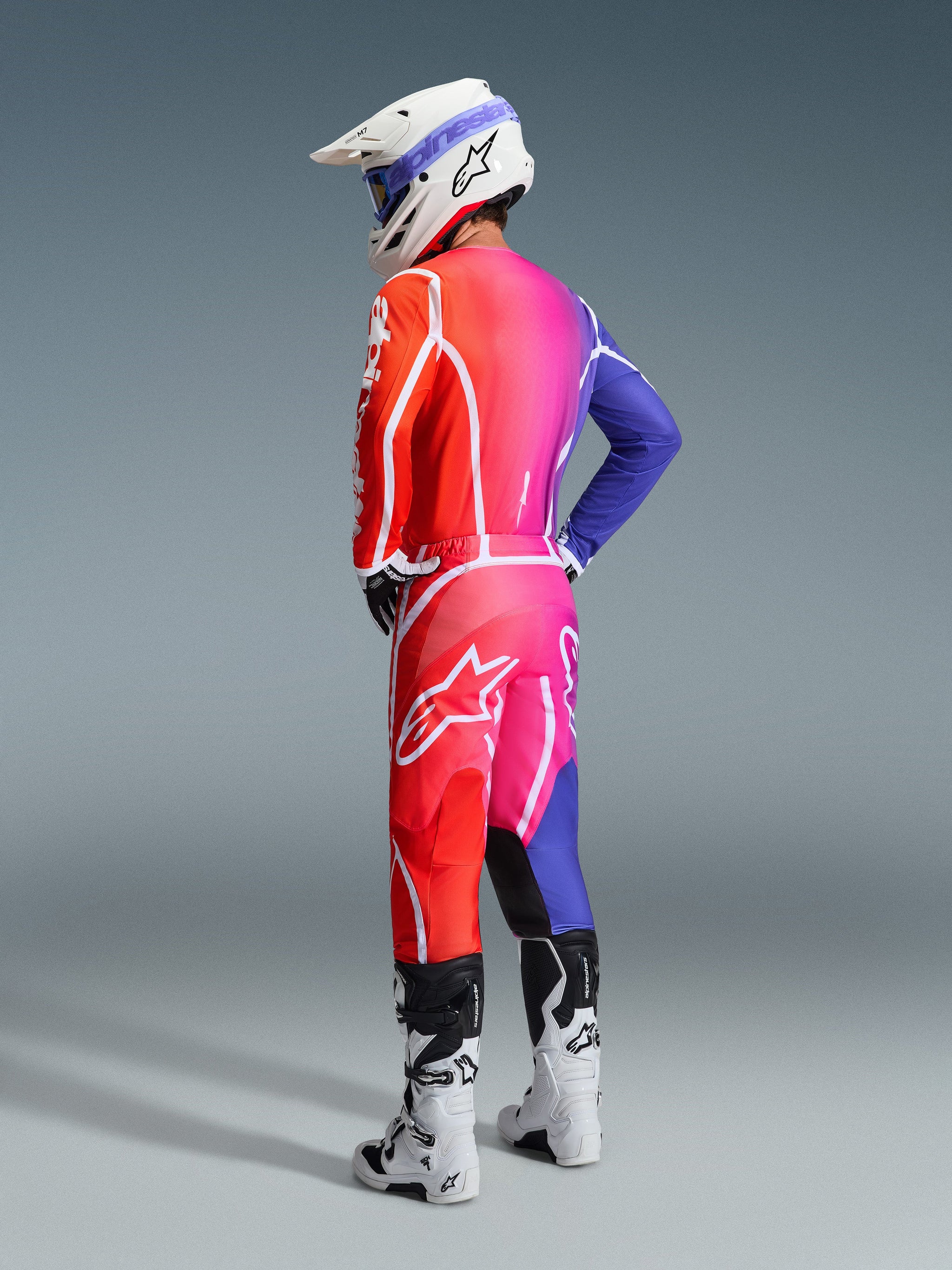 Motocross-kører iført Alpinestars 2026 Fluid Apex Jersey og matchende bukser, levende rød, pink og blå gradient med hvide detaljer, hvid hjelm med blå briller og hvide støvler, set bagfra af komplet off-road racerudstyr.