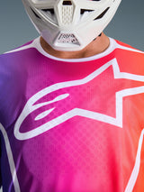 Alpinestars 2026 Fluid Apex trøje, motocross-trøje, flerfarvet lilla, lyserød og orange gradient, med et stort hvidt Astars-logo på brystet, nærbillede båret med en hvid motocrosshjelm