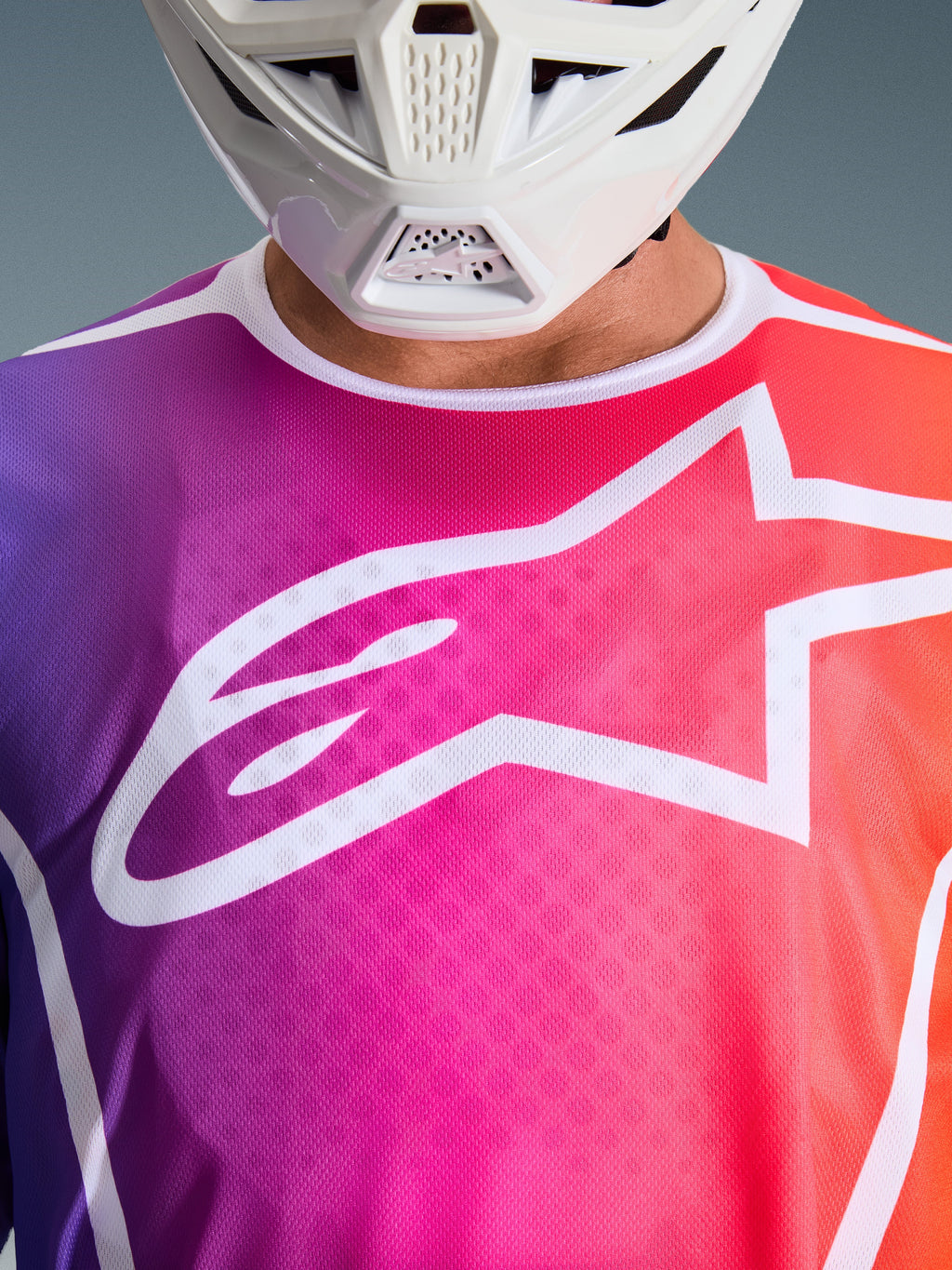 Alpinestars 2026 Fluid Apex trøje, motocross-trøje, lilla, pink og orange farveovergang, stort hvidt Alpinestars-stjernelogo på brystet, nærbillede der viser hvid hagebeskytter fra hjelm