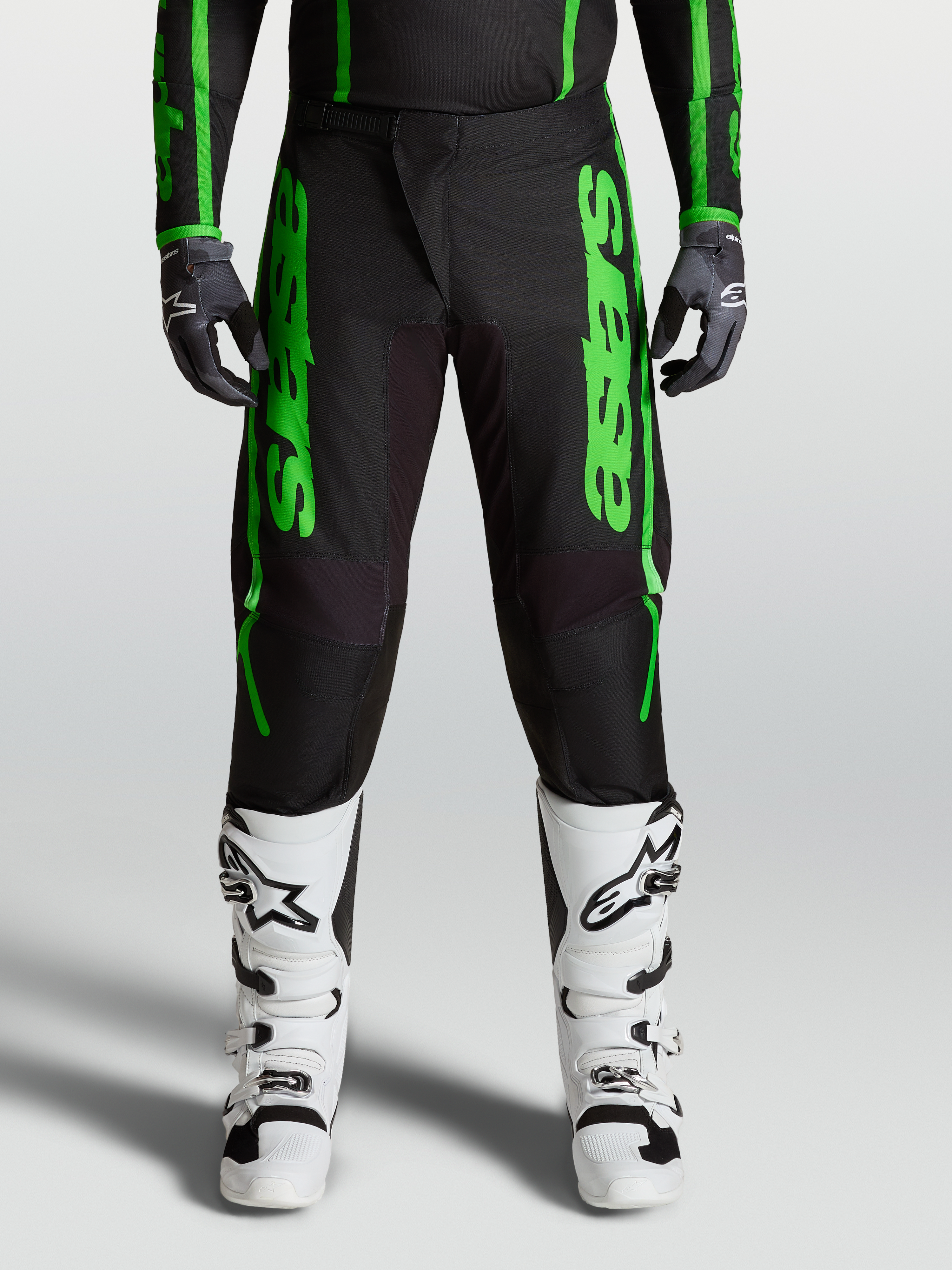 Alpinestars Fluid Apex V2 bukser, motocross-bukser i grøn og sort, forstærkede knæ og polyfabric-konstruktion, båret med hvide støvler, avanceret off-road ydeevne.
