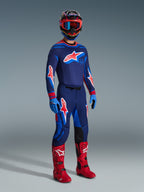 Motocross-kører i komplet racerudstyr med Alpinestars 2026 Supertech Vista Jersey i Dark Navy, Hot Coral og Cobalt, matchende bukser med markante stjernelogoer, klare røde motocross-støvler, blå handsker og en flerfarvet hjelm med røde goggles, professionelt udstyrssæt til terrænkørsel.