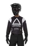 Alpinestars Techdura Jersey, Black, featuring a large white triangle chest graphic with ASTARS logo and white shoulder panels, worn by a rider with a black motocross helmet, goggles, and matching black off-road gear.