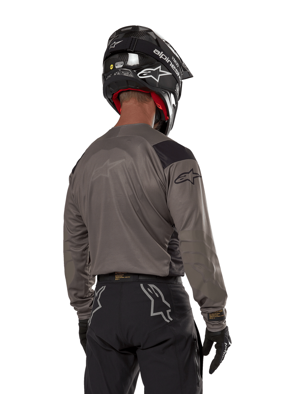 Motocross rider from the back wearing the Alpinestars Techdura Jersey in Falcon Brown, paired with black off-road pants and a black helmet, featuring a modern graphic design and elongated rear for enduro riding
