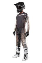 Motocross-rytter iført komplet racerudstyr, 2024 Techstar Pneuma Jersey i Dark Sand og Iron Dust Gray, matchende mønstrede motocross-bukser, sort hjelm med goggles, hvide og sorte støvler, sorte handsker, koordineret sæt til off-road racing