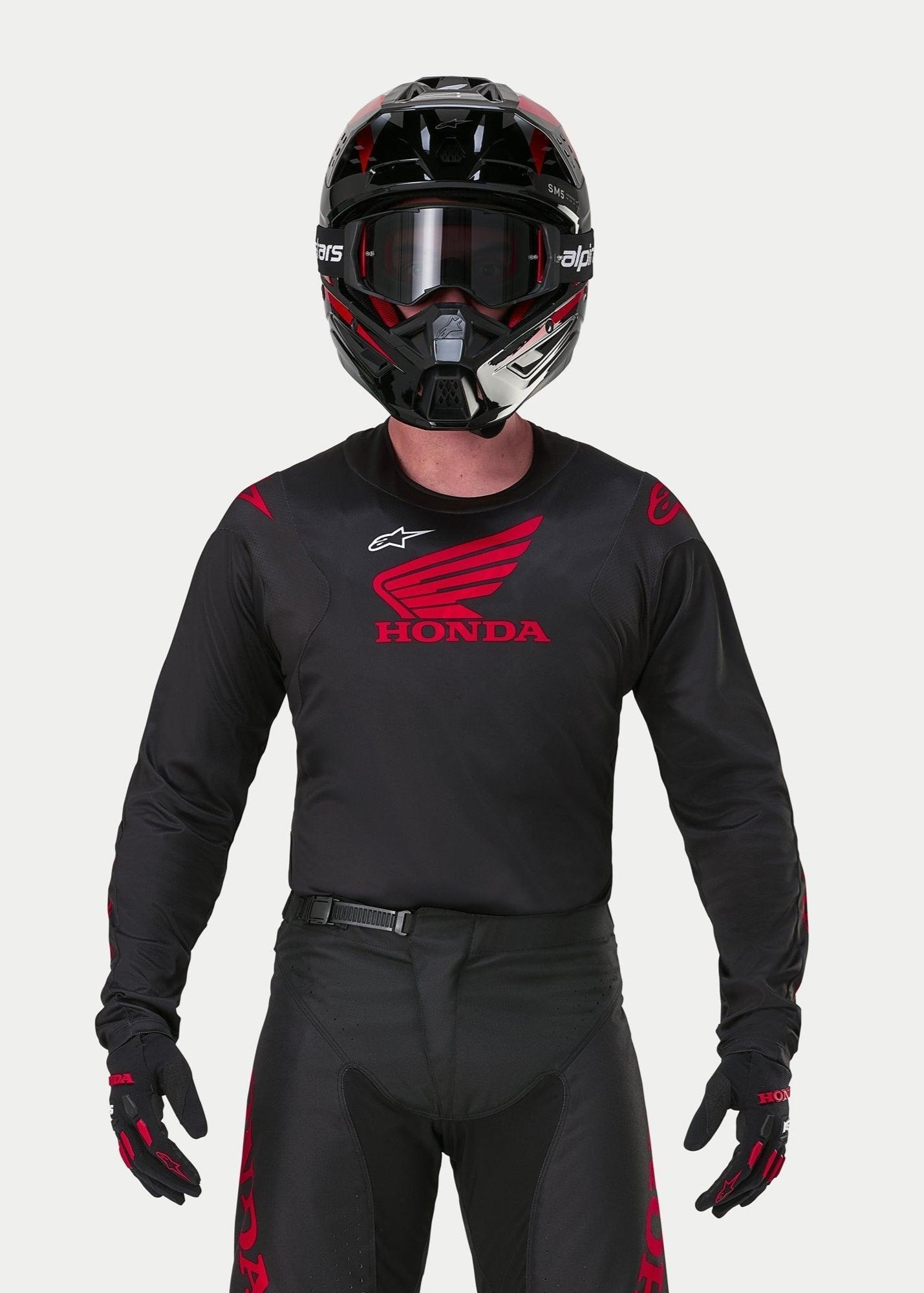 Motocrosskører iført Alpinestars Honda Racer Iconic Jersey i sort og rød, med et stort rødt Honda-vingelogo på brystet og hvidt Astar-logo, kombineret med matchende sort hjelm, briller og handsker for et komplet off-road racersæt