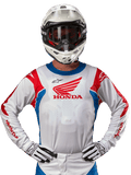 Honda Racer Iconic Jersey, hvid motocross-trøje med rødt Honda-logo og klare blå og røde detaljer, båret af en kører med hvid hjelm og handsker, højtydende udstyr til off-road og motocross-racing