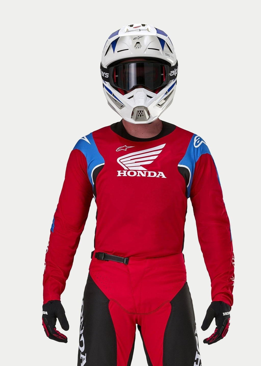 Motocrosskører iført Alpinestars Honda Racer Iconic trøje, klar rød med blå og hvide detaljer og Honda-branding, kombineret med en hvid og blå hjelm, mørke goggles og matchende røde og sorte motocrossbukser, præstationsudstyr til terrænkørsel