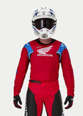 Motocrosskører iført Alpinestars Honda Racer Iconic trøje, klar rød med blå og hvide detaljer og Honda-branding, kombineret med en hvid og blå hjelm, mørke goggles og matchende røde og sorte motocrossbukser, præstationsudstyr til terrænkørsel