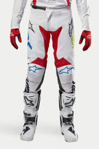 Alpinestars 2024 Racer Hana bukser, motocross-bukser, hvid multicolor med røde, blå og limegrønne detaljer, båret med hvide og sorte støvler og røde handsker, med Alpinestars-logoer på knæene og et holdbart teknisk design til off-road racing