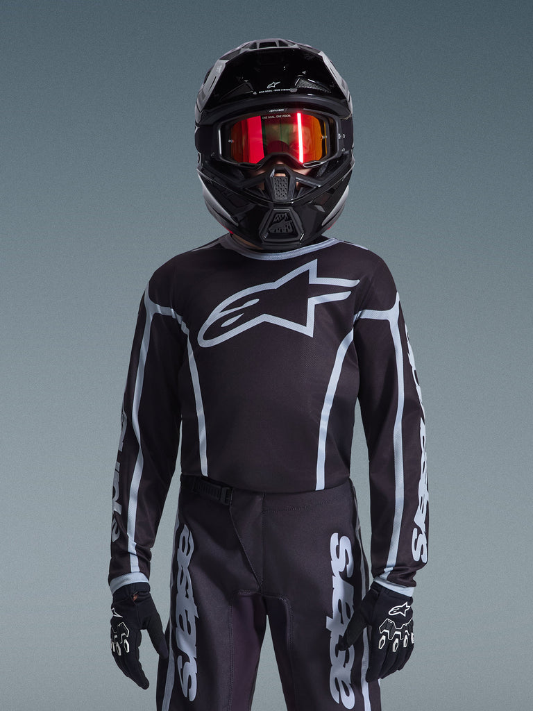 Youth motocross rider wearing Alpinestars 2026 Youth Fluid Apex Jersey in Black and Gray, matching pants, black helmet with red reflective goggles, and black gloves, coordinated off-road racing gear.
