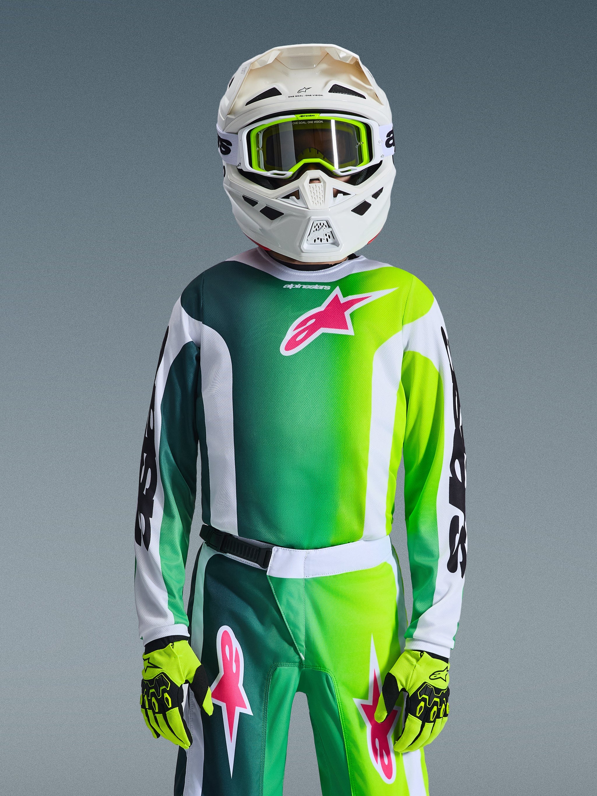 Ung motocross-rytter iført komplet racerudstyr, grøn, hvid og sort 2026 Youth Fluid Portl trøje med pink logo-accent, matchende bukser og handsker, hvid motocross-hjelm med lime-goggles, koordineret outfit til off-road kørsel