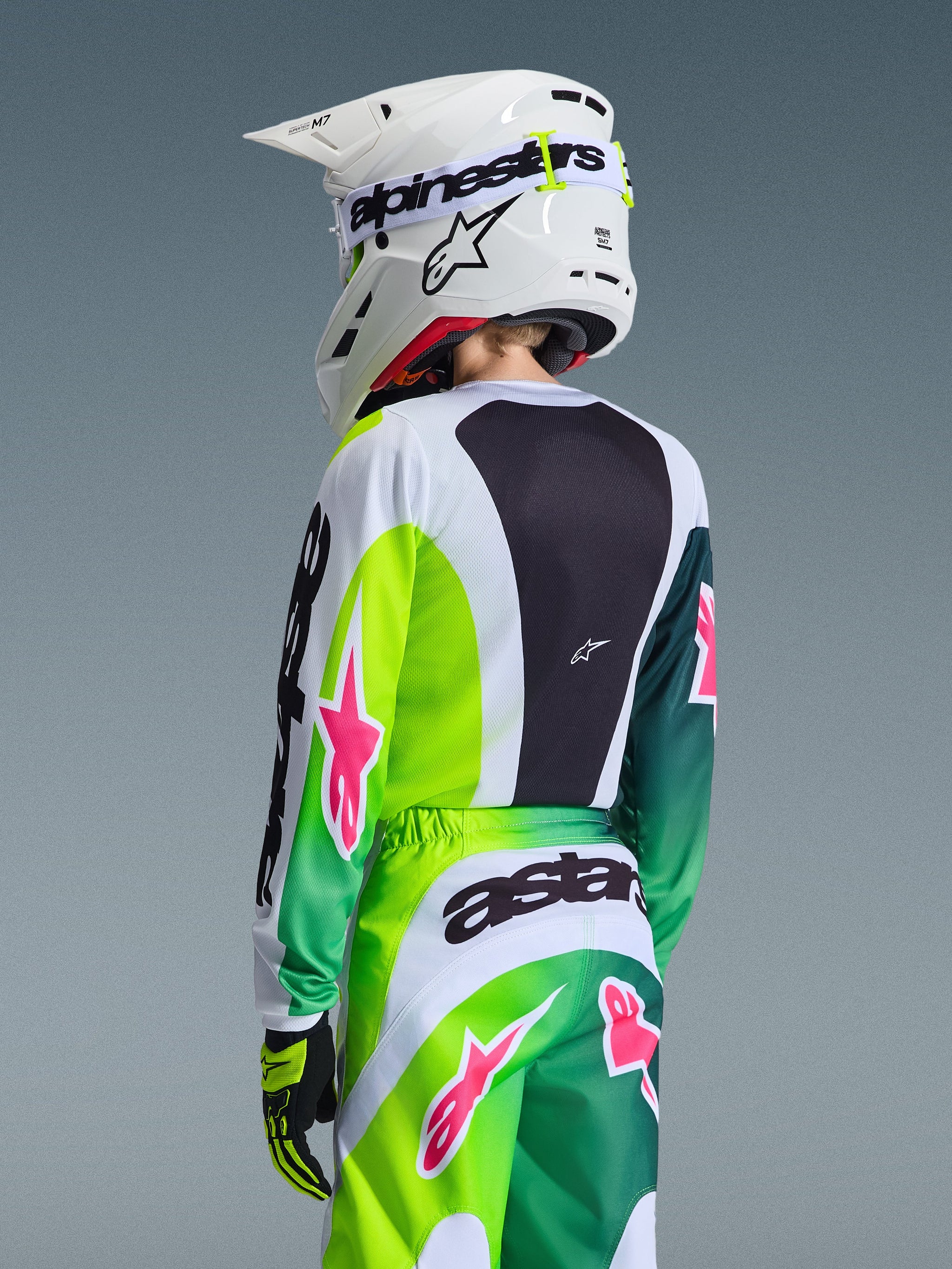 Alpinestars 2026 Youth Fluid Portl trøje, grøn hvid og sort med lyserøde stjerne-accenter, set bagfra af en ung rytter iført matchende motocross-udstyr og en hvid Alpinestars-hjelm, sportsligt ergonomisk design til off-road kørsel