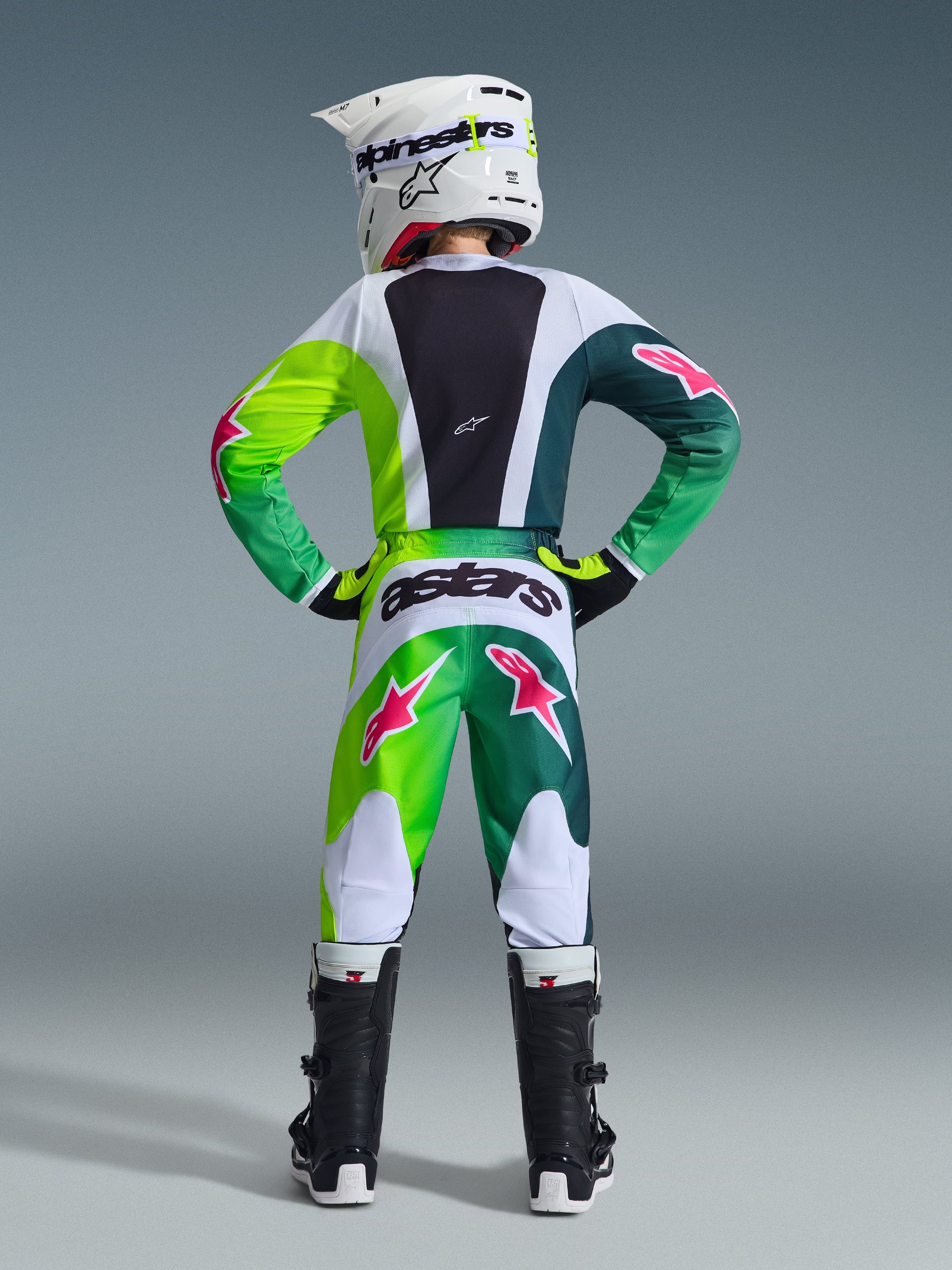 Alpinestars 2026 Youth Fluid Portl trøje, grøn, hvid og sort, ungdomsmotocrossrytter vist bagfra iført matchende bukser med lyserøde stjernelogoer, hvid hjelm og sorte støvler, koordineret præstationsudstyr til off-road kørsel.