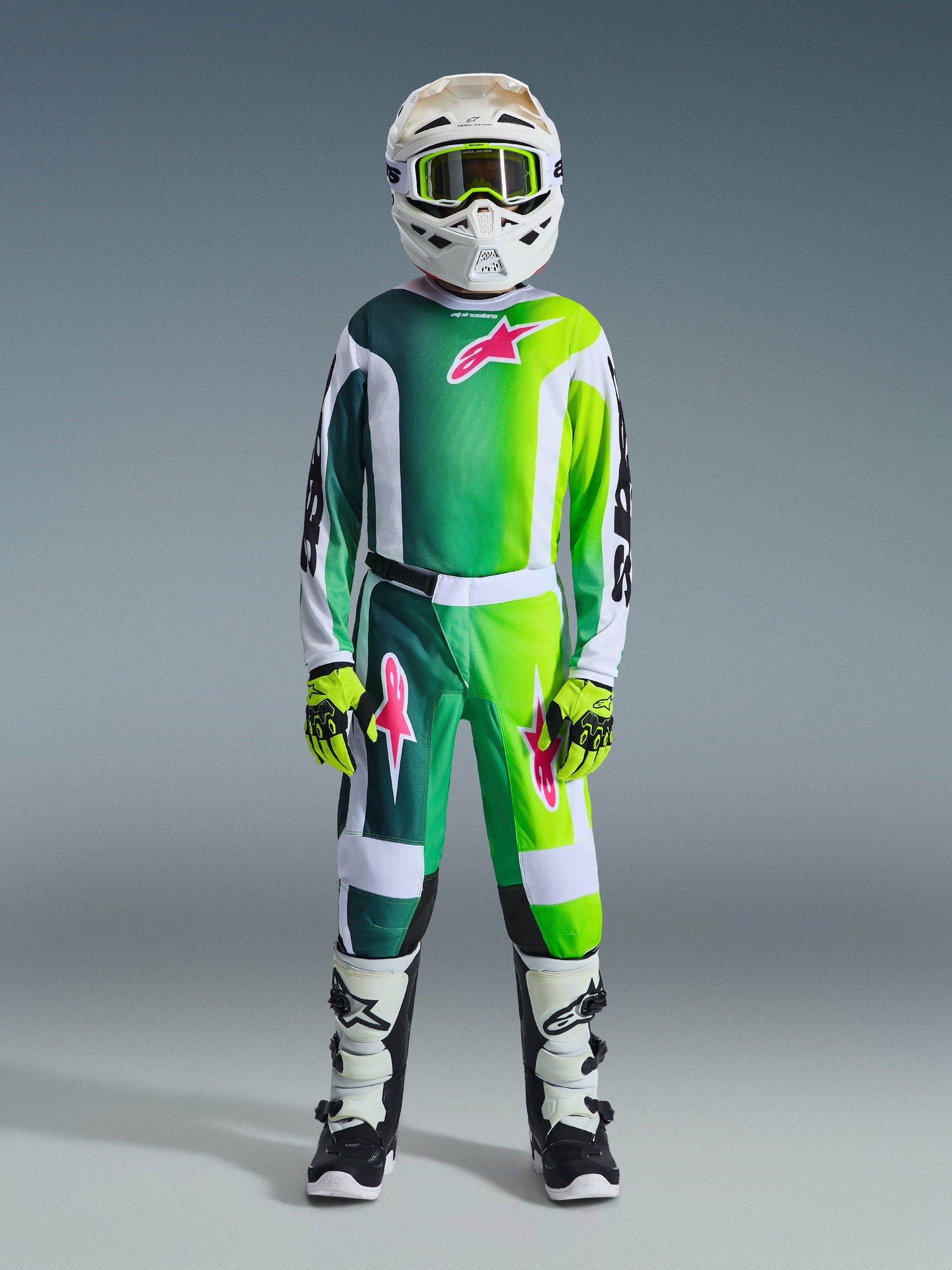 Motocross-rytter iført komplet racerudstyr, 2026 Youth Fluid Portl Jersey i grøn, hvid og sort med lyserødt logo, matchende bukser, hvid hjelm med gule briller, hvide og sorte støvler, limegrønne handsker, koordineret sæt til off-road kørsel for unge