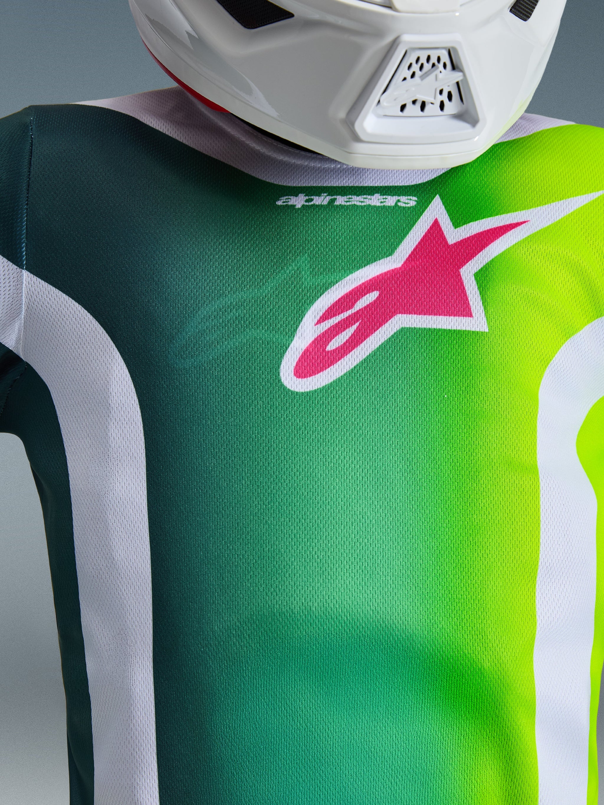 Alpinestars 2026 Youth Fluid Portl Jersey, motocross-trøje, grøn og hvid med et stort lyserødt Alpinestars-logo på brystet, nærbillede af kraven og teknisk mesh-stof, båret med en hvid hjelm