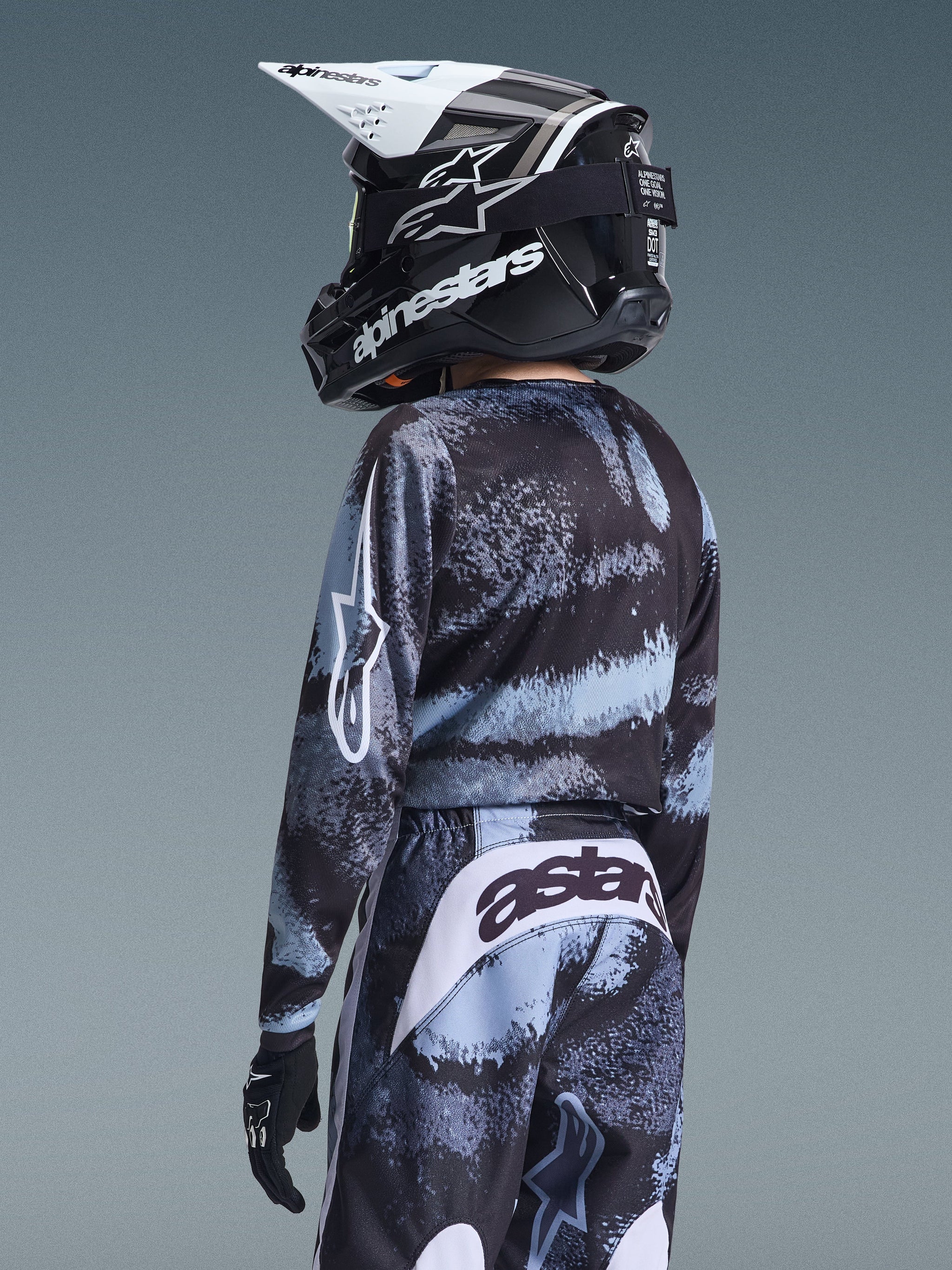 Youth rider wearing complete motocross gear from behind, Alpinestars 2026 Youth Fluid Lahnd Jersey in Iron Camo with black and light blue patterns, matching pants with 'astars' logo on the seat, black and white helmet, and black gloves.