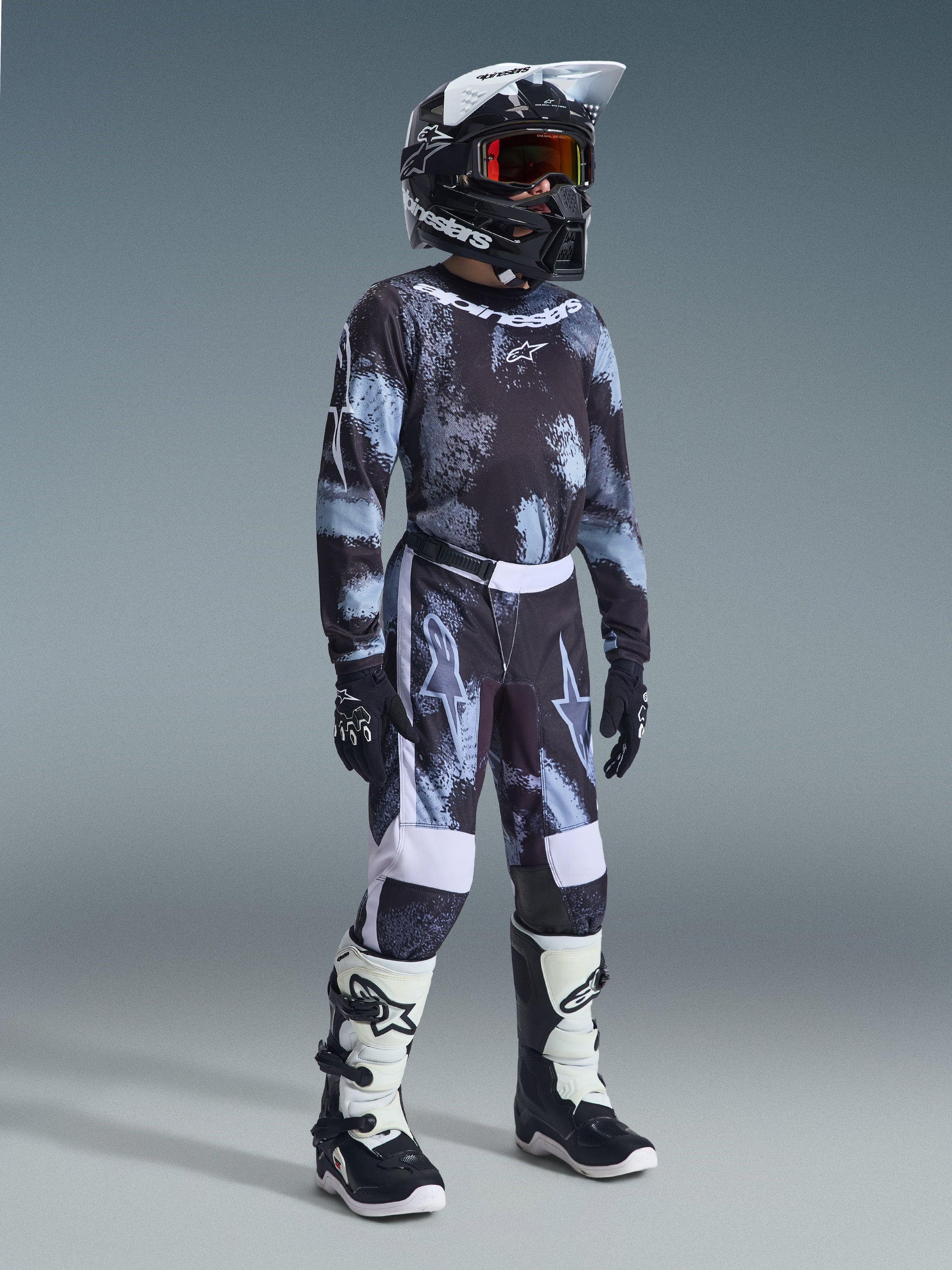 Youth motocross rider wearing the 2026 Youth Fluid Lahnd Jersey in Iron Camo, black and grey camouflage pattern, matching pants, black Alpinestars helmet with goggles, black and white boots, and black gloves, complete youth motocross racing gear