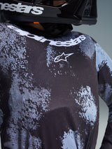Close-up of the Alpinestars 2026 Youth Fluid Lahnd Jersey in Iron Camo, featuring a black and grey camouflage pattern, white Alpinestars logo on the chest, and brand name along the neckline, worn by a rider in a black helmet.