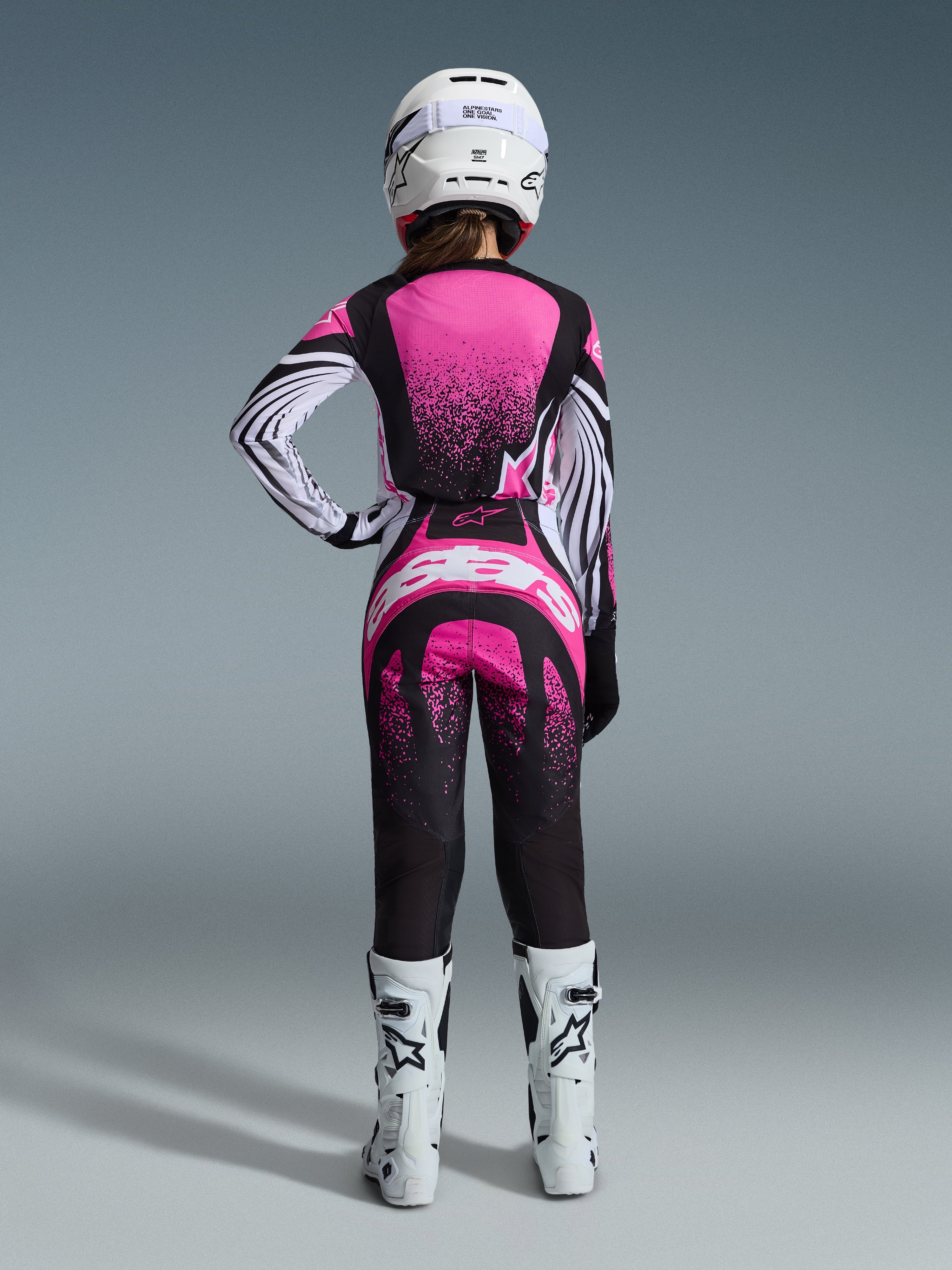 Alpinestars 2026 Stella Techstar Nomur-udstyrssæt, kvindelig motocrosskører set bagfra, lyserød og sort jersey med hvidstribede ærmer og nistret mønster, matchende bukser med hvidt Alpinestars-logo, hvid hjelm og hvide off-road støvler